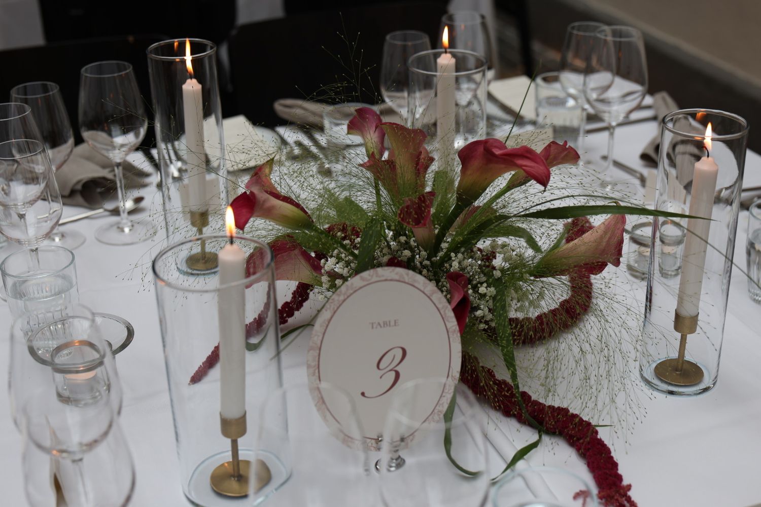 Elegant wedding table centerpiece with white taper candles, red calla lilies, and a table number 3 card on white linen.