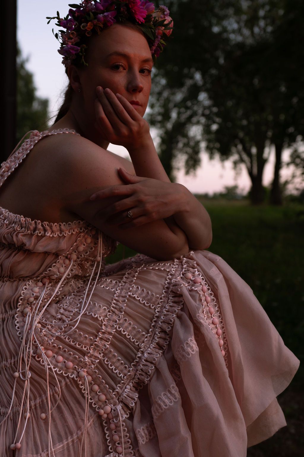 Woman in pink embroidered dress and floral crown poses thoughtfully outdoors at dusk near trees.