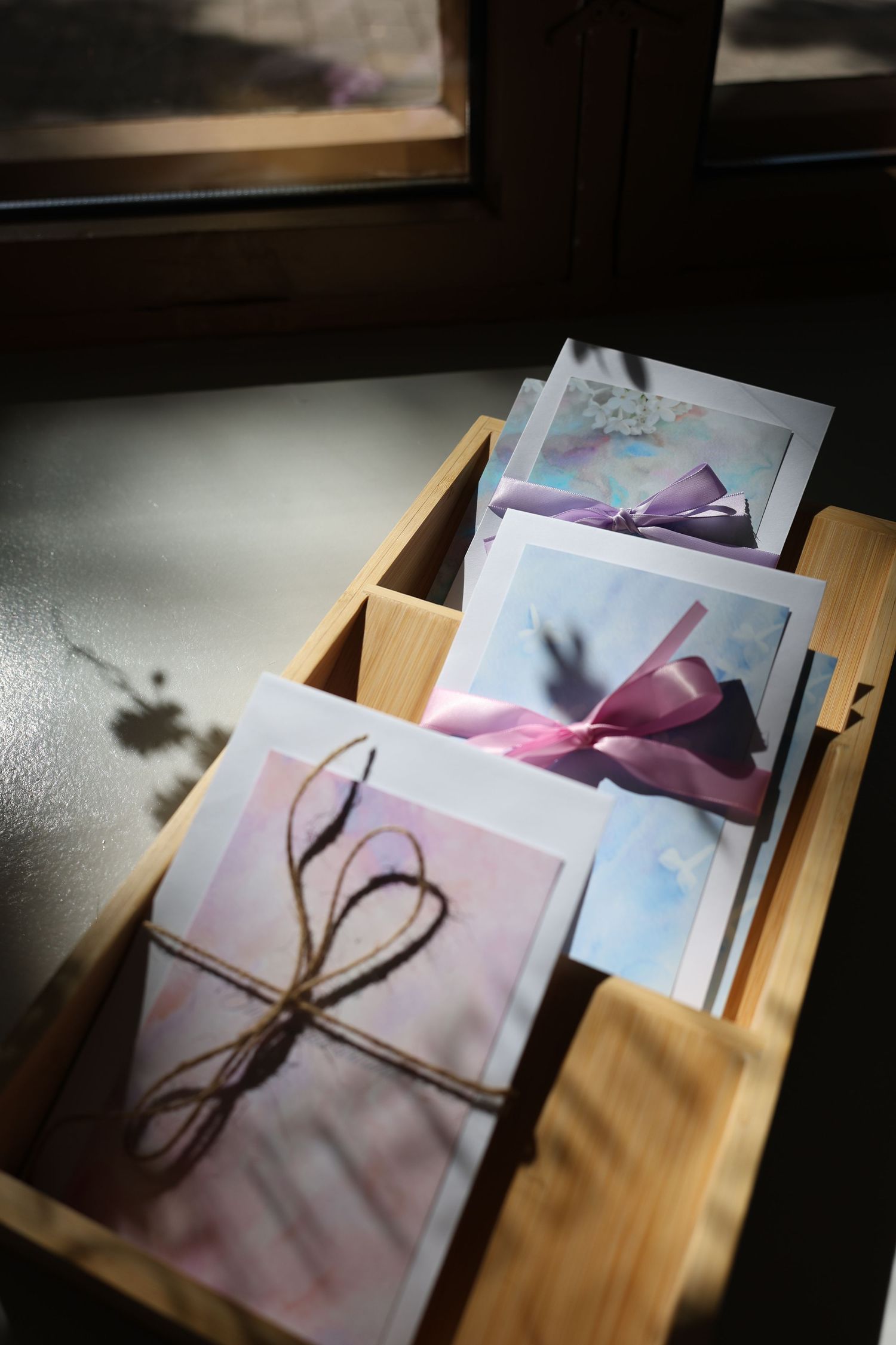 Wooden box with greeting cards tied with twine and pink ribbon bows, displayed near a sunlit window.
