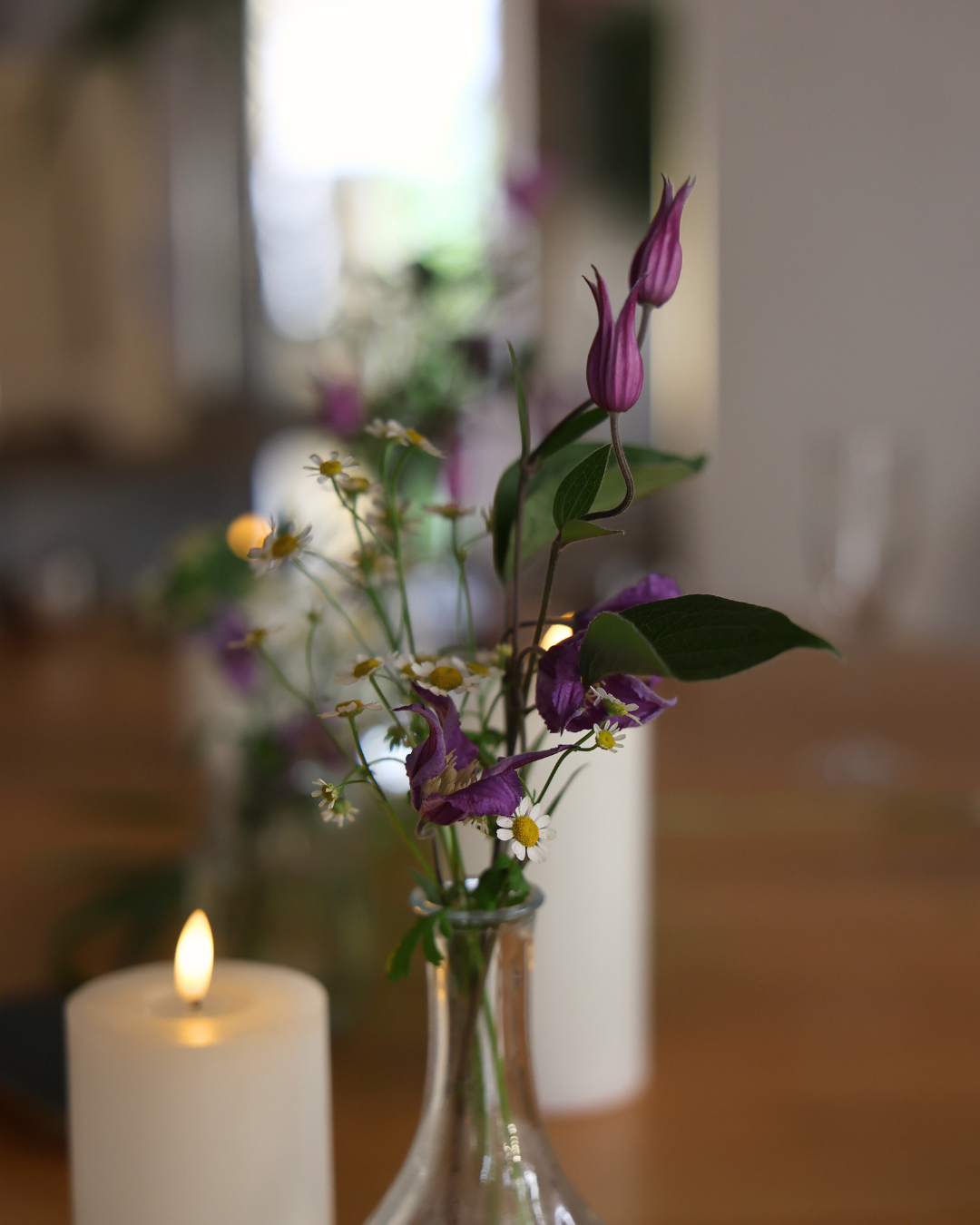 Glass vase with purple tulips and white wildflowers beside a lit candle on a wooden table.