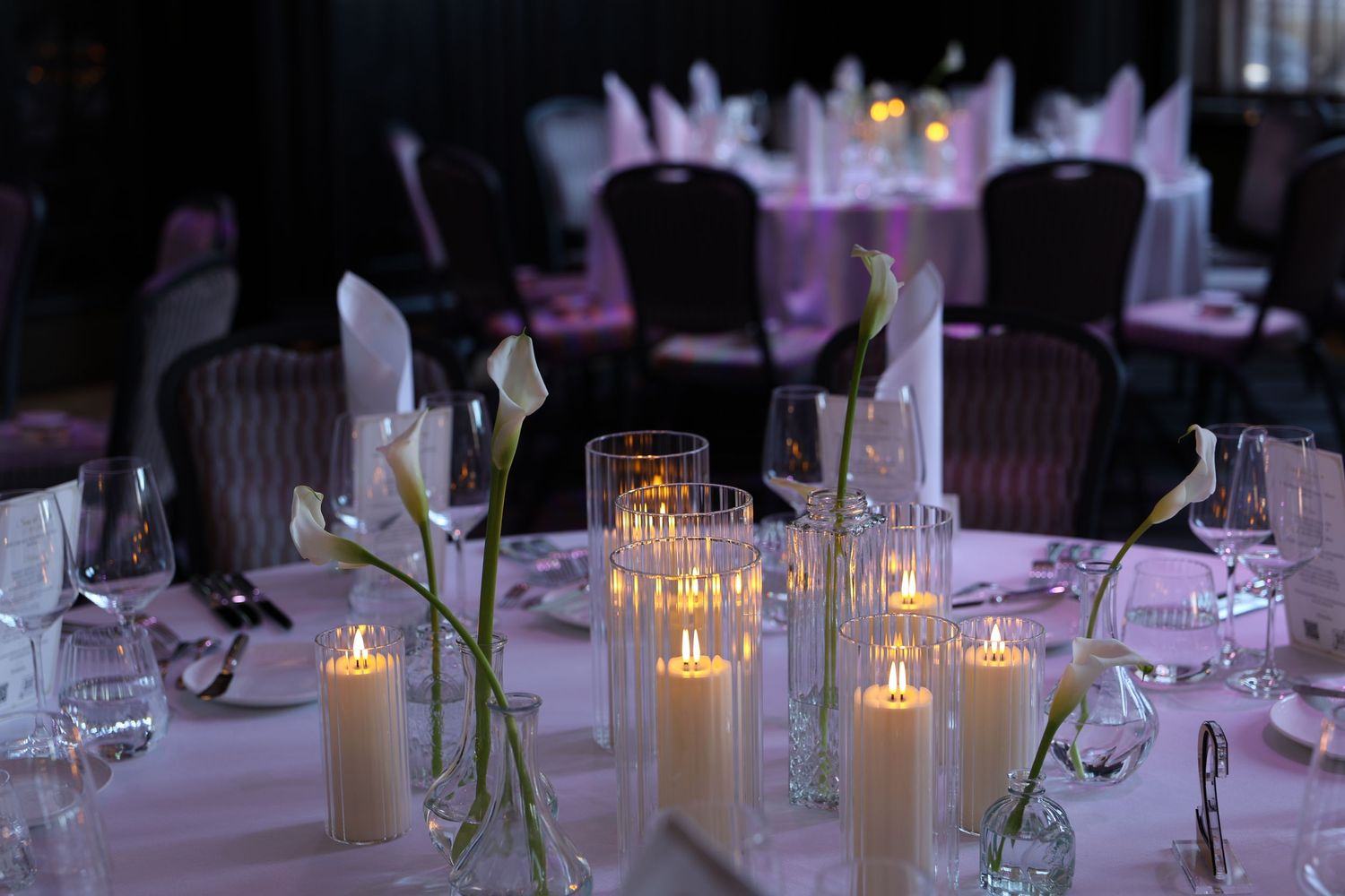 Elegant wedding reception table with white pillar candles, calla lilies, and crystal glassware on pink linen.