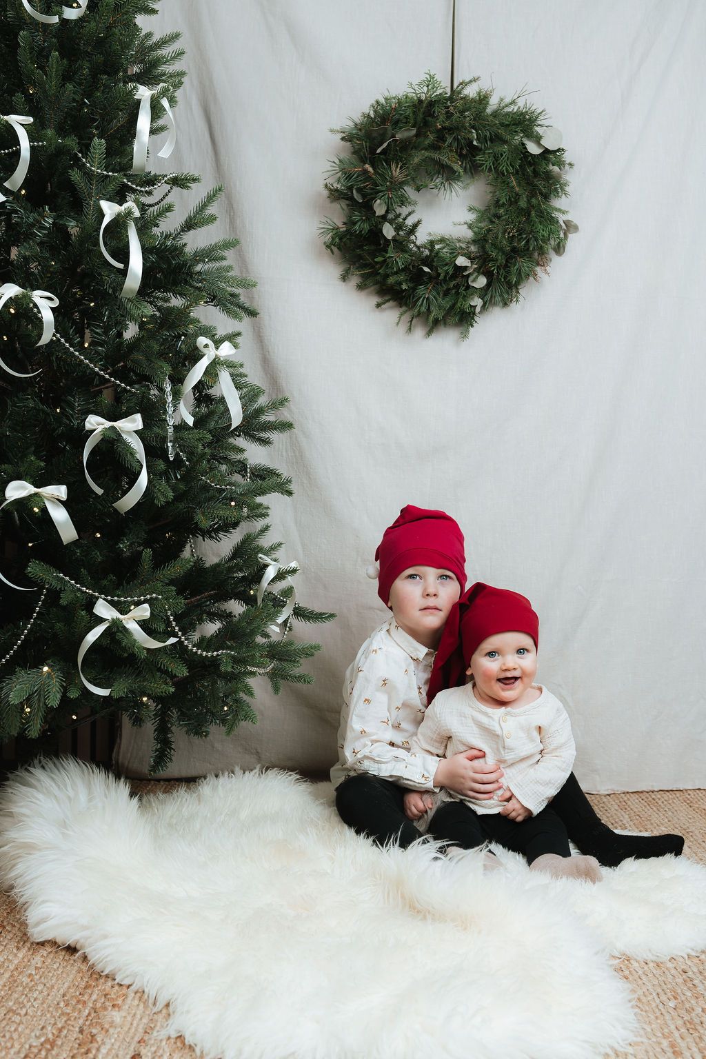 Two children in red Santa hats sit on a white fur rug beside a decorated Christmas tree with a wreath backdrop.
