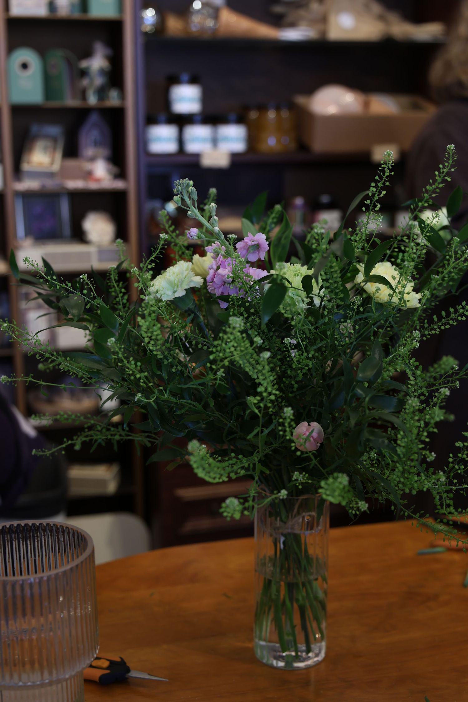 A fresh floral arrangement with green foliage and purple flowers in a glass vase sits on a wooden table with shelving in background.