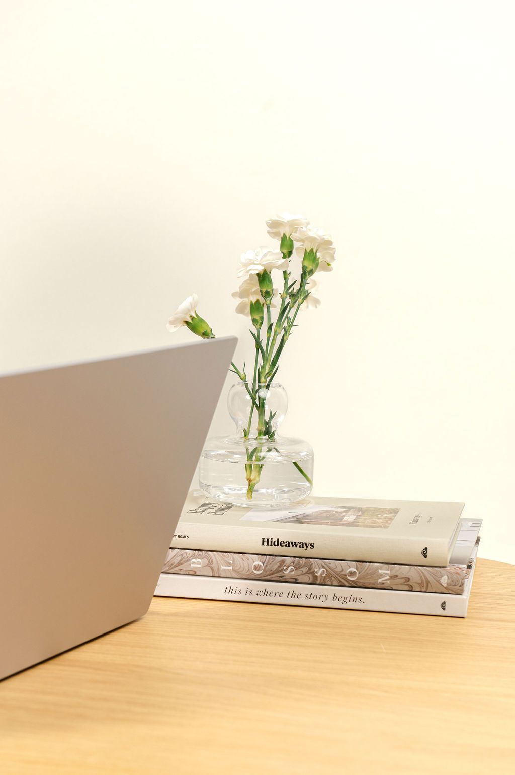 A minimalist desk setup with stacked books, a glass vase of white flowers, and a laptop on a wooden surface.