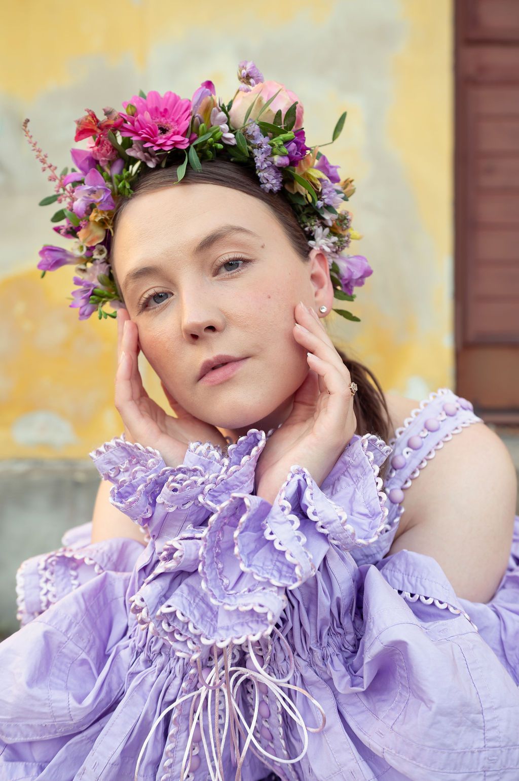 Ethereal spring portrait featuring floral crown with pink dahlias and purple blooms atop lavender ruffled dress.