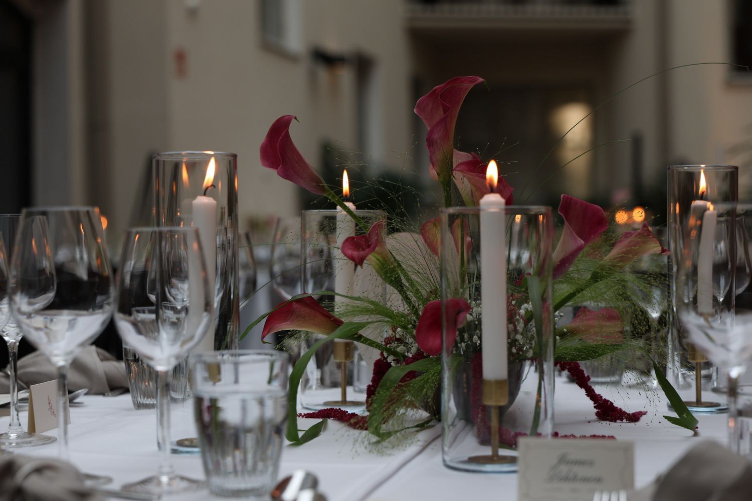 Elegant wedding reception table with red calla lilies, lit candles, and crystal glassware on a white tablecloth.