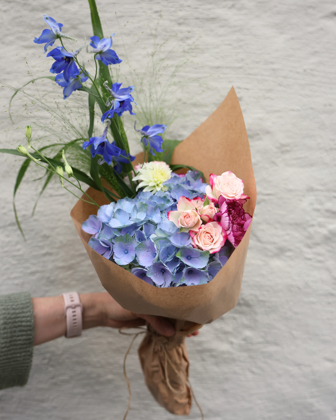 Hand holding a kraft paper-wrapped bouquet with blue hydrangeas, pink roses, and delphinium against a white wall.