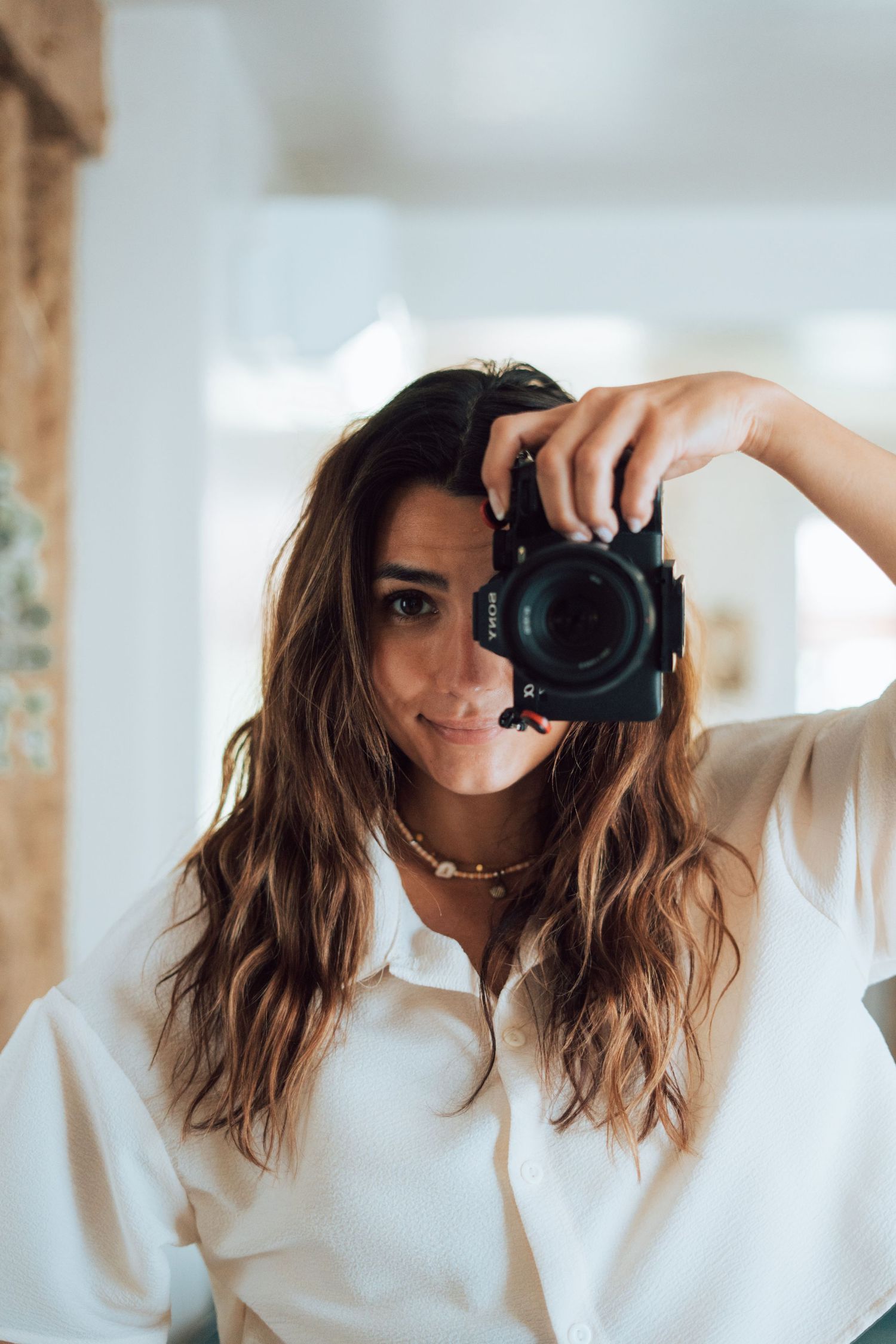 Woman in white shirt holding a DSLR camera up to her face, smiling, with long wavy brown hair indoors.