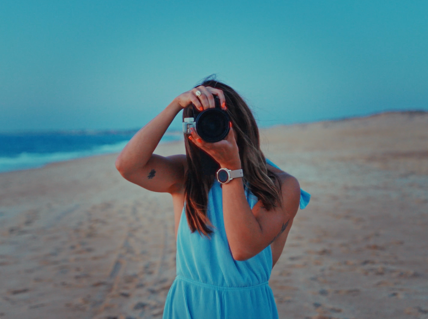 Woman in blue dress photographing on a sandy beach with ocean in background on a clear day.