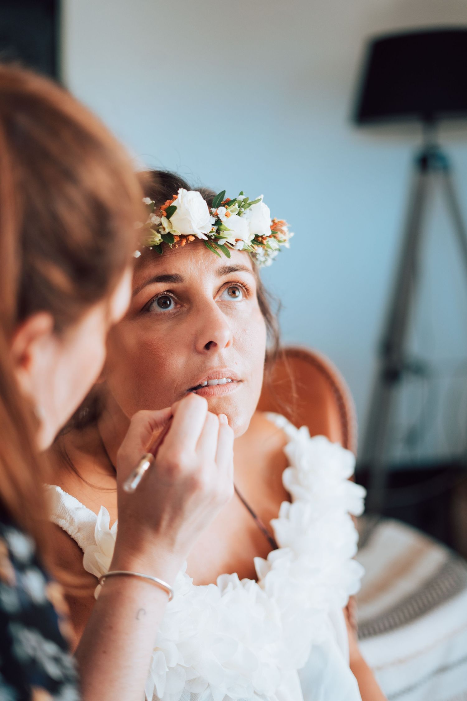 Bride wearing flower crown gets makeup touch-up from bridesmaid before wedding ceremony.
