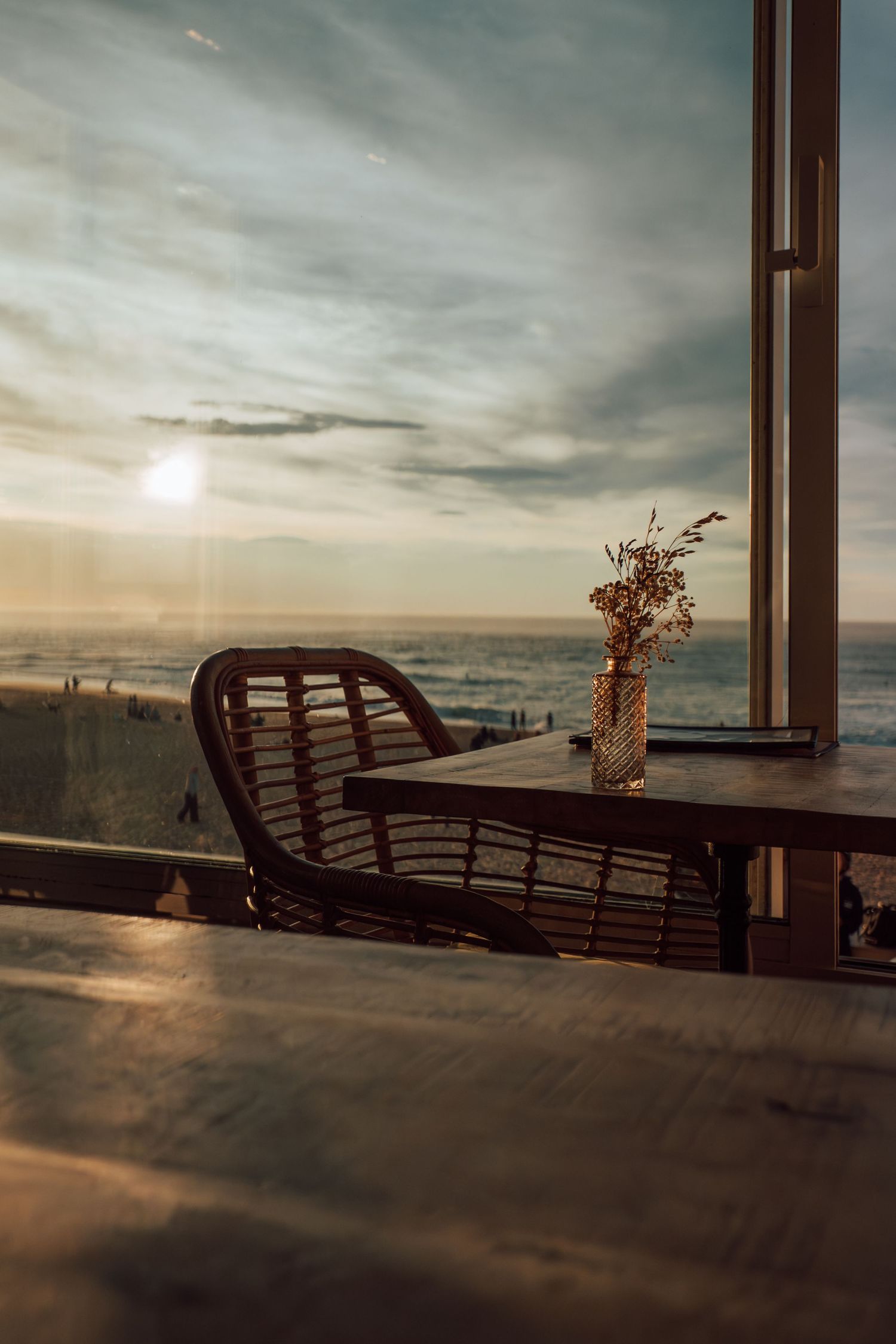 Wicker chair and vase with dried flowers on a wooden table overlooking a stunning ocean sunset through large windows.