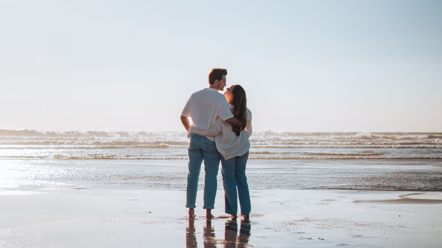 Romantic couple standing together on a beach, embracing while looking at the ocean on a sunny day.
