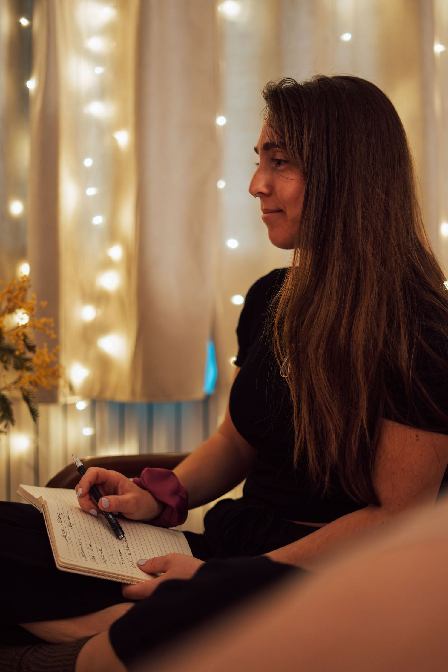 Woman with long hair writing in notebook, sitting in cozy room with warm fairy lights in background.