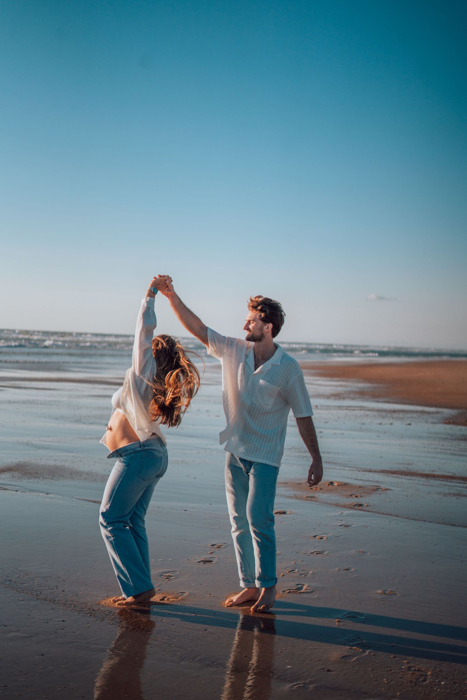 Joyful couple dancing barefoot on a wet beach at sunset, man spinning woman with raised hands.