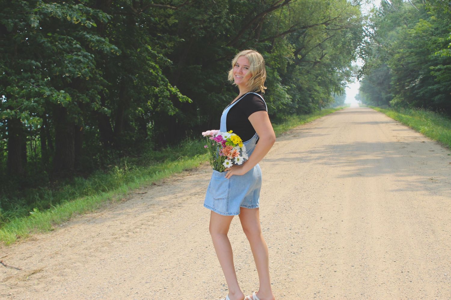 Blonde woman holds wildflowers on a sunny tree-lined dirt road.