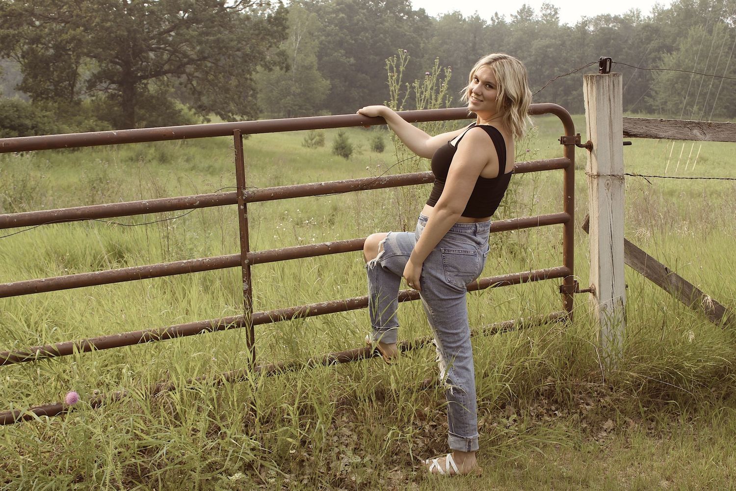Blonde woman poses against rustic fence in green countryside field.