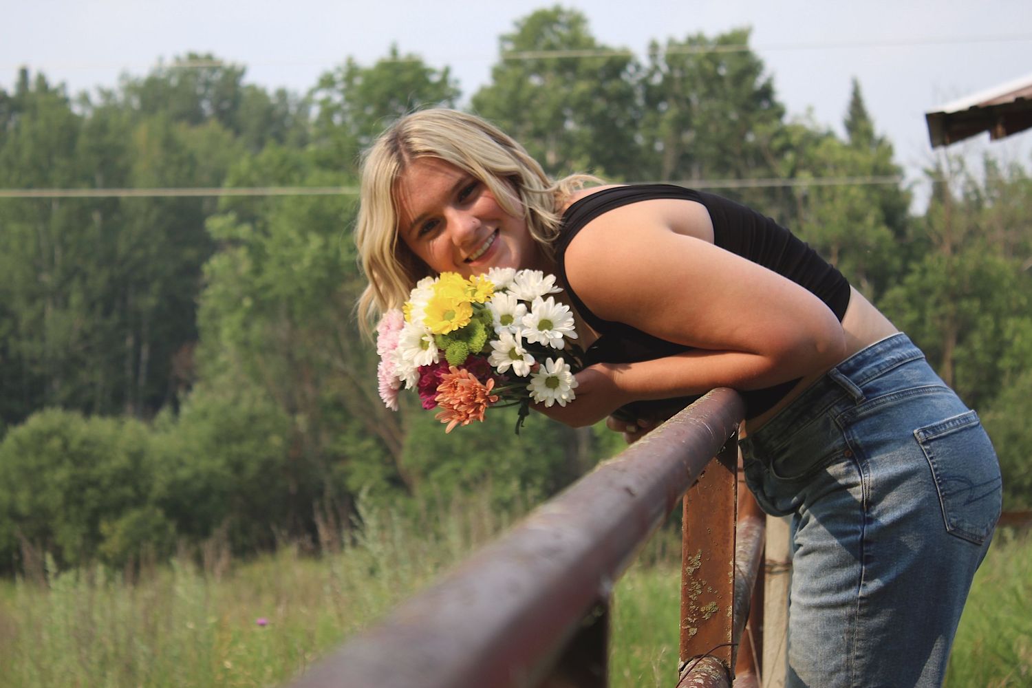 A smiling blonde woman leans over a wooden fence holding a colorful bouquet of flowers.