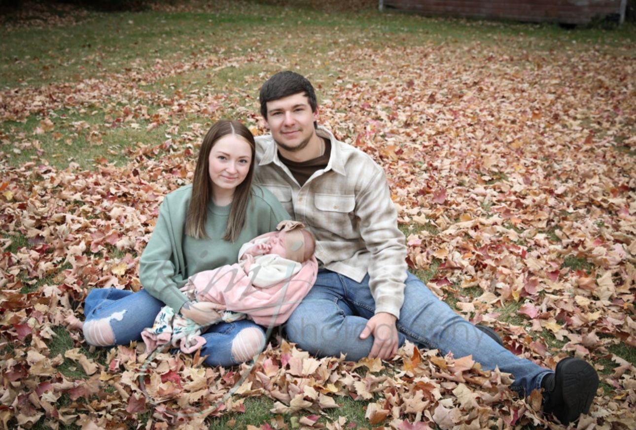 A young couple sits together on a bed of autumn leaves holding their newborn baby.
