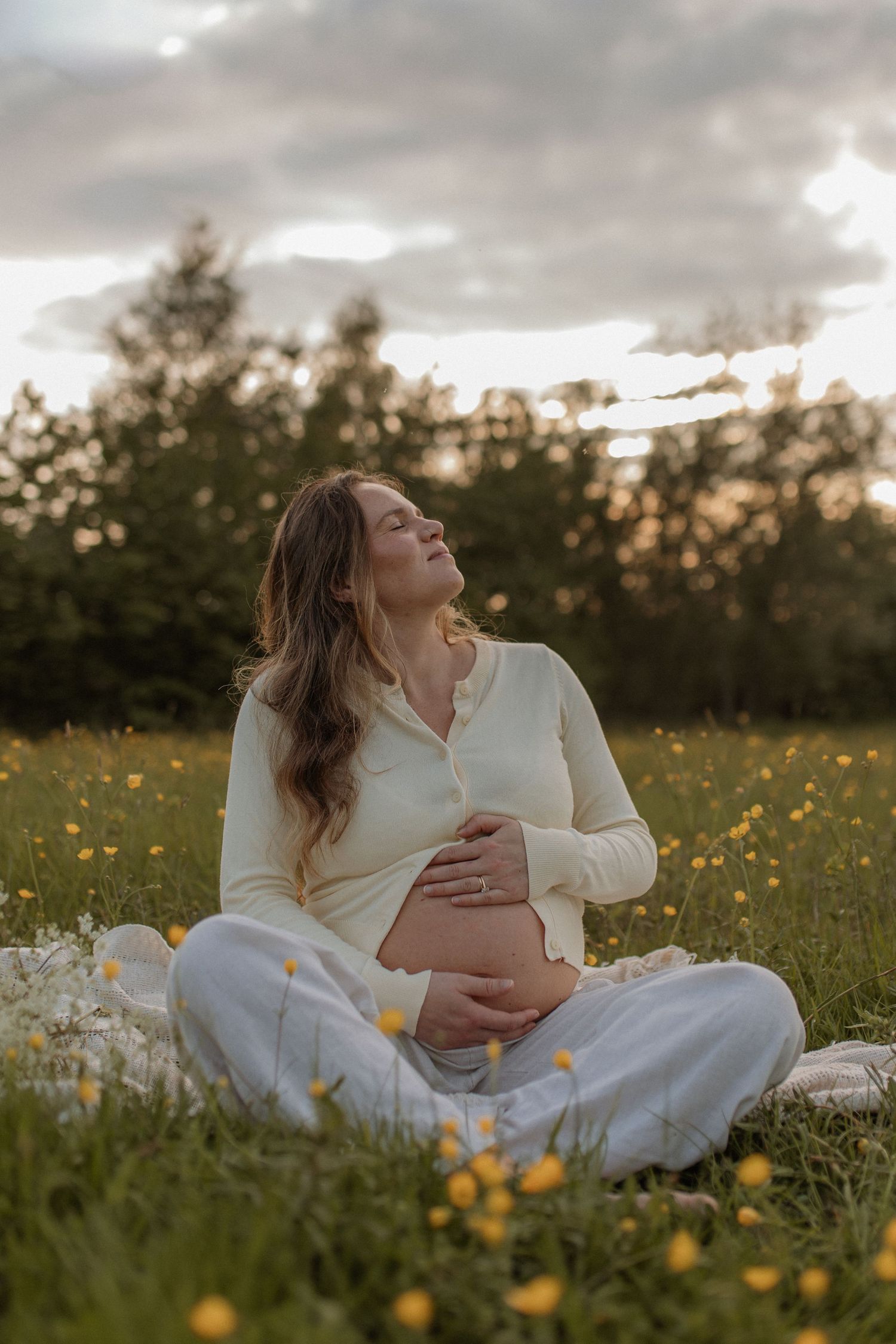 Pregnant woman in white outfit sitting in flower field at sunset, holding baby bump peacefully.