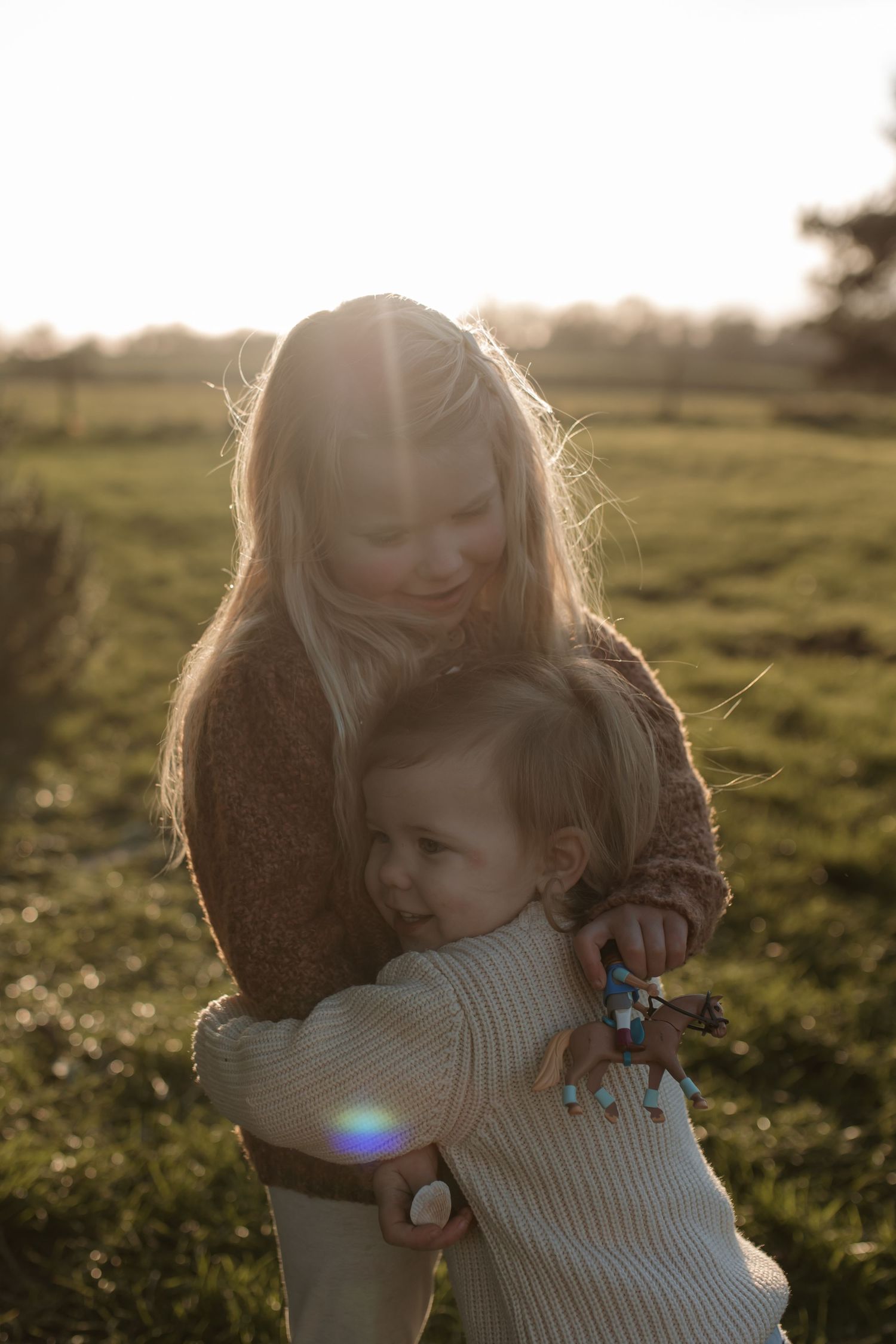 Older sister lovingly hugging young toddler sibling in a sun-drenched green field.