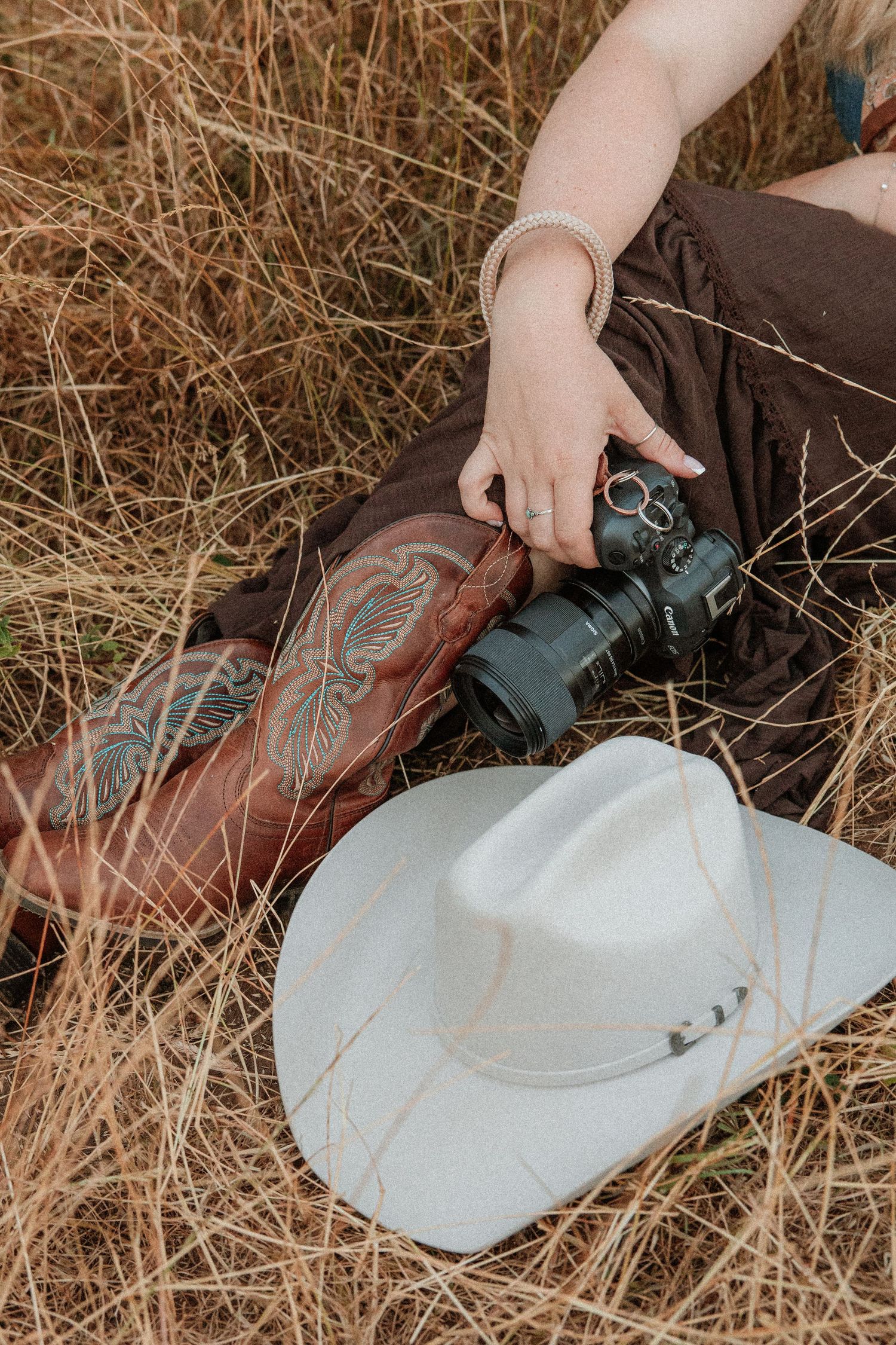 Woman in brown skirt holding camera beside cowboy boots and white hat in dry grass field.