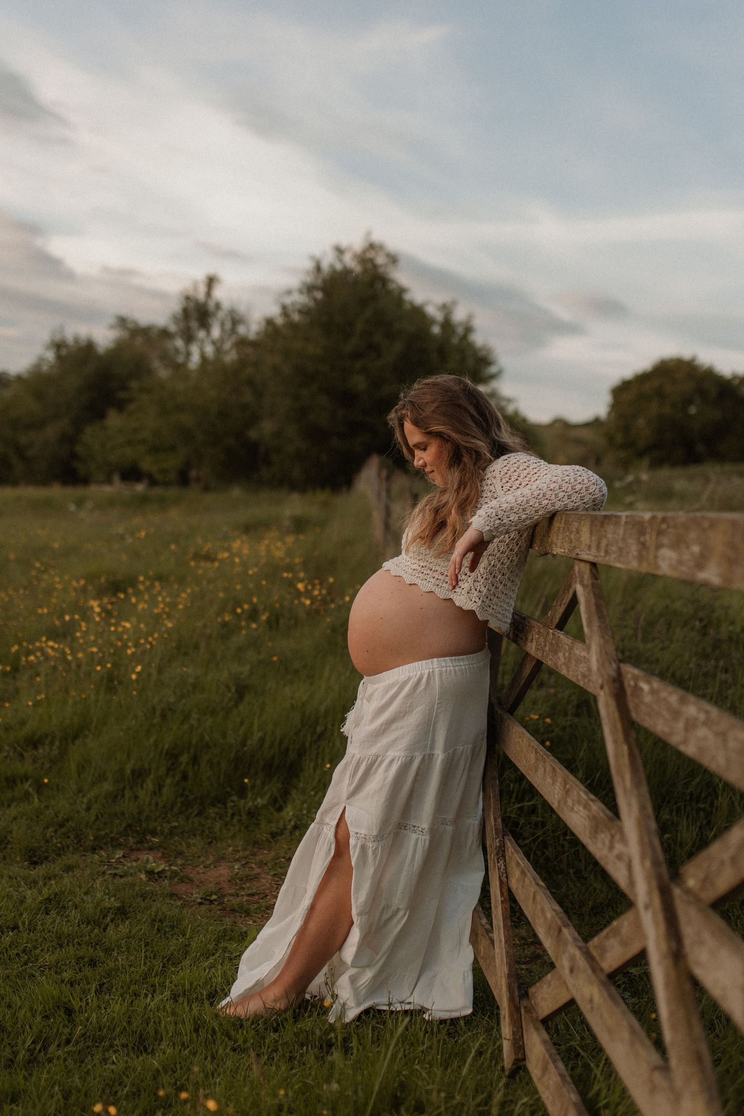 A pregnant woman in a white skirt and crochet top leans against a wooden fence in a golden meadow.