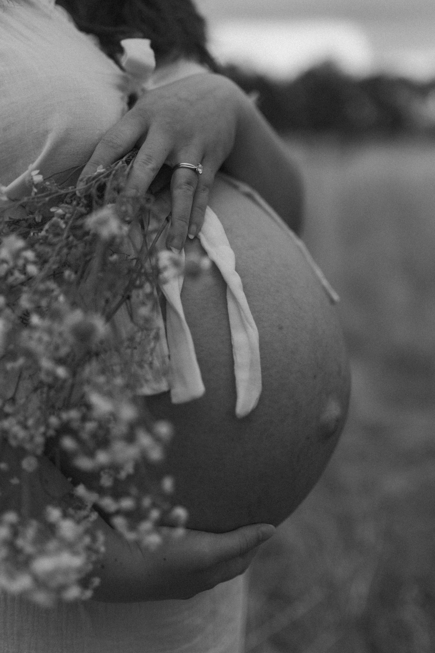 Black and white photo of pregnant belly with hand holding wildflowers in field at dusk.
