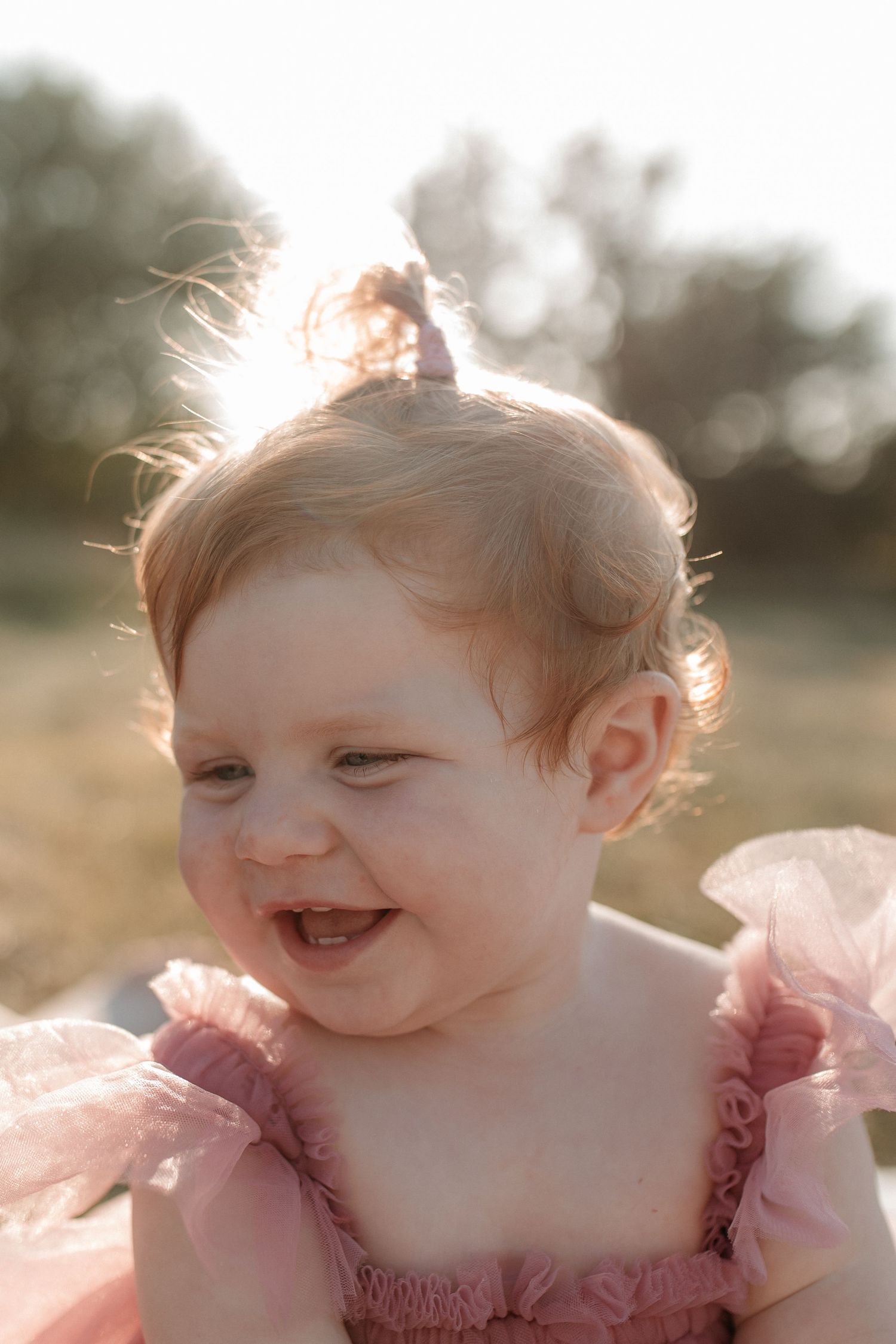 Happy red-haired baby girl in pink ruffle dress laughing in golden sunlight outdoors.