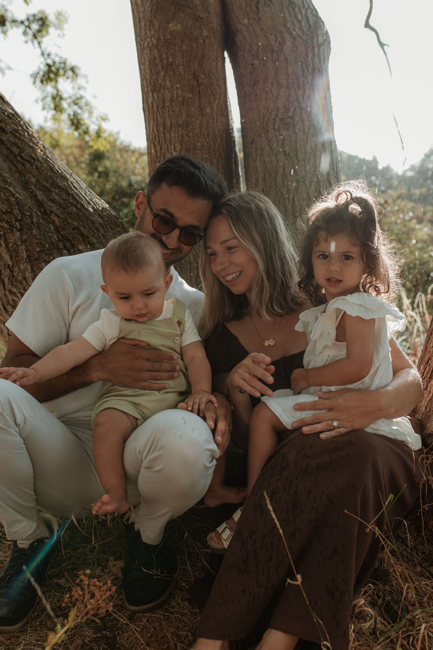 A happy family of four sits together under trees in warm sunlight.