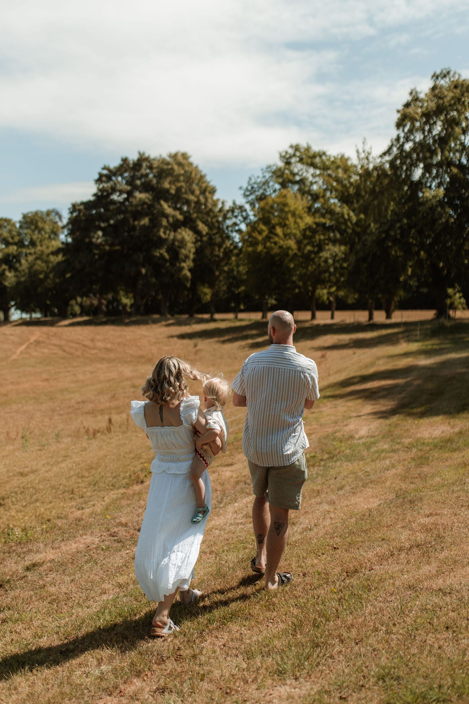 A couple walks through a sunny open field carrying their young baby.
