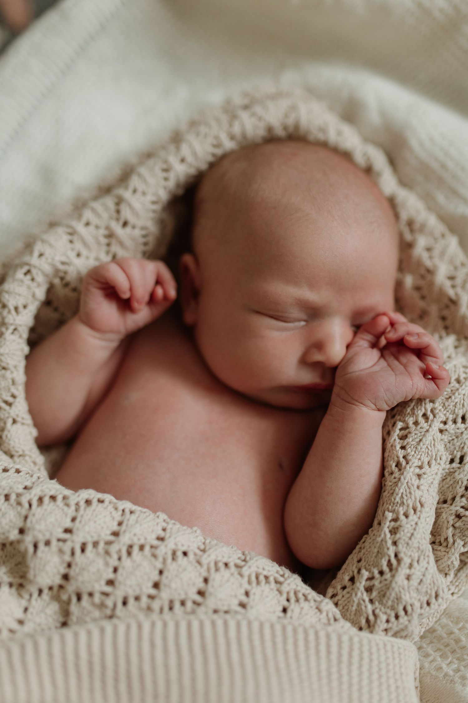 Newborn baby sleeping peacefully wrapped in a soft cream knitted blanket with hands raised.