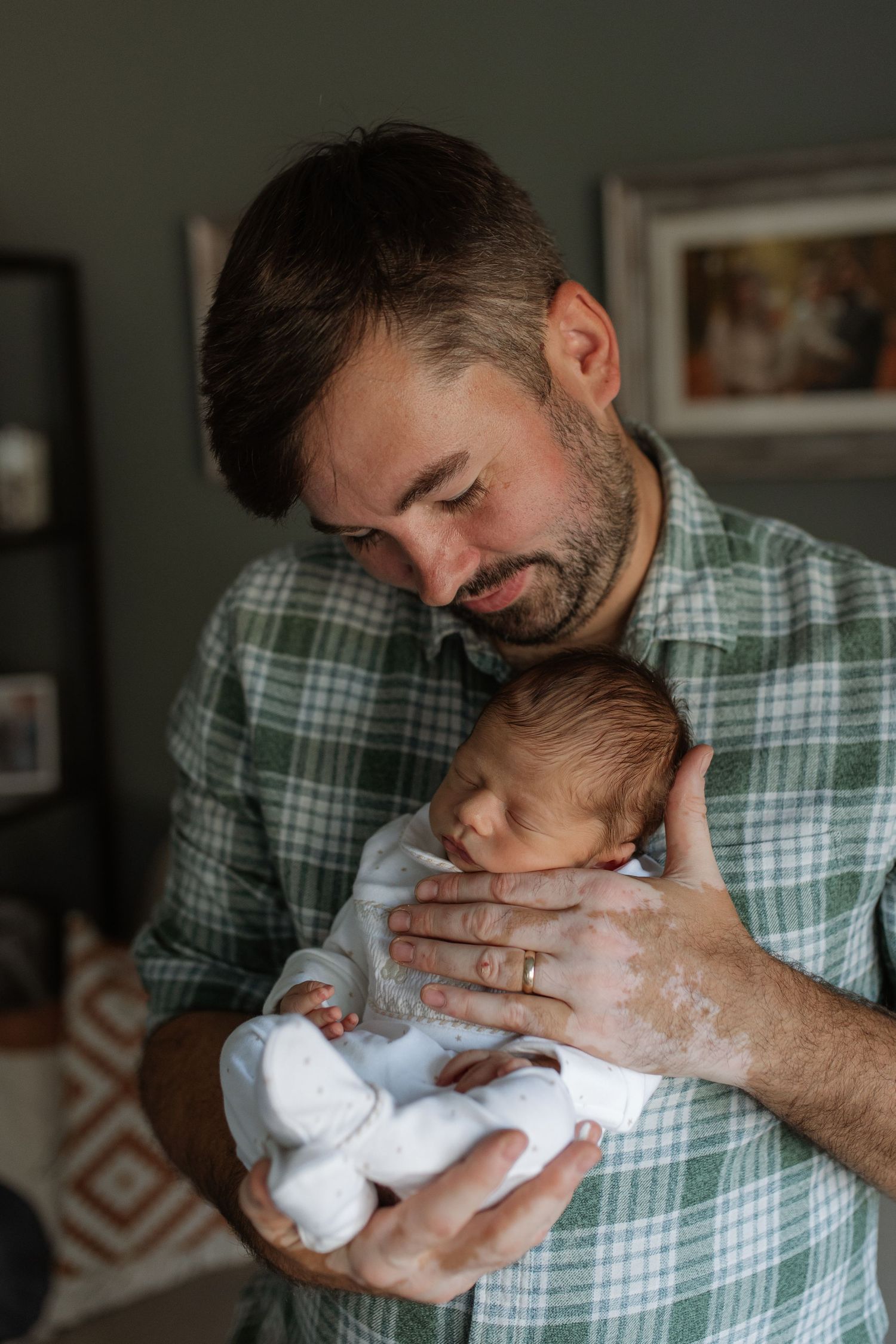 Loving father in green plaid shirt cradling sleeping newborn baby against his chest.