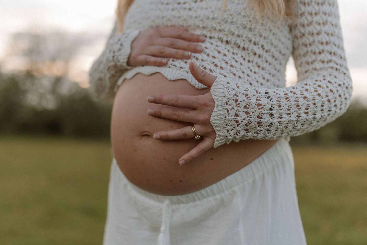 Close-up of a pregnant woman cradling her bare baby bump with both hands wearing a lace sleeve top.