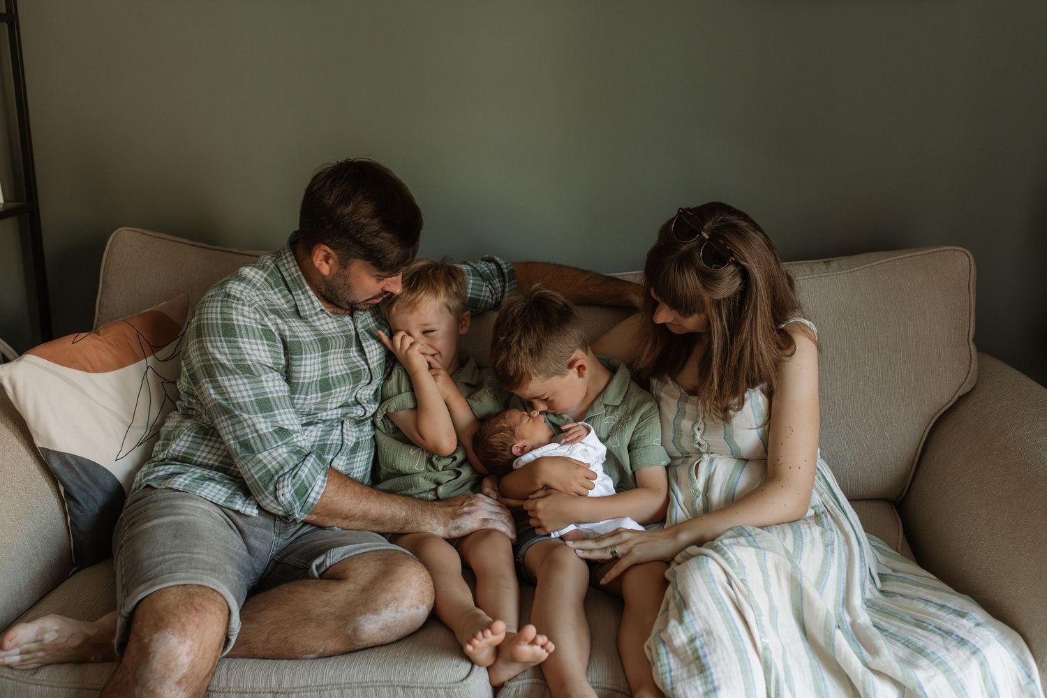Family of five gathered on a couch admiring their newborn baby together indoors.