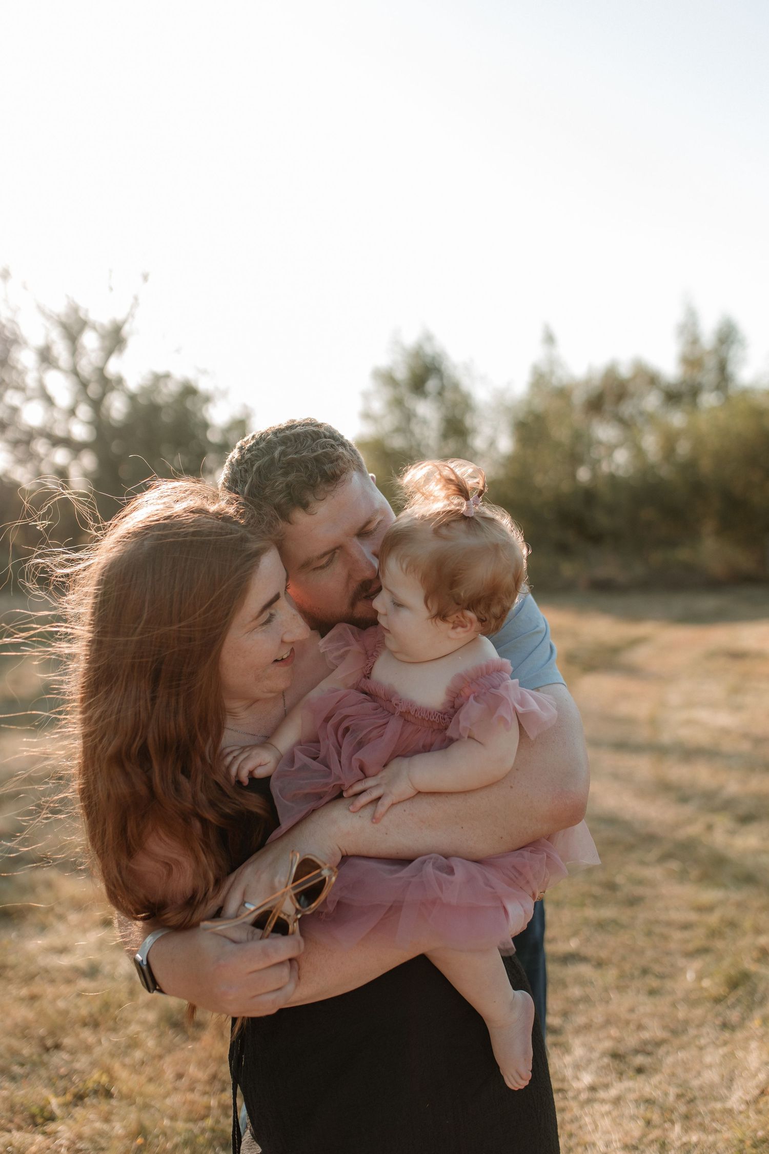 A family of three sharing a tender moment outdoors, parents kissing their baby girl in a pink tutu dress at golden hour.