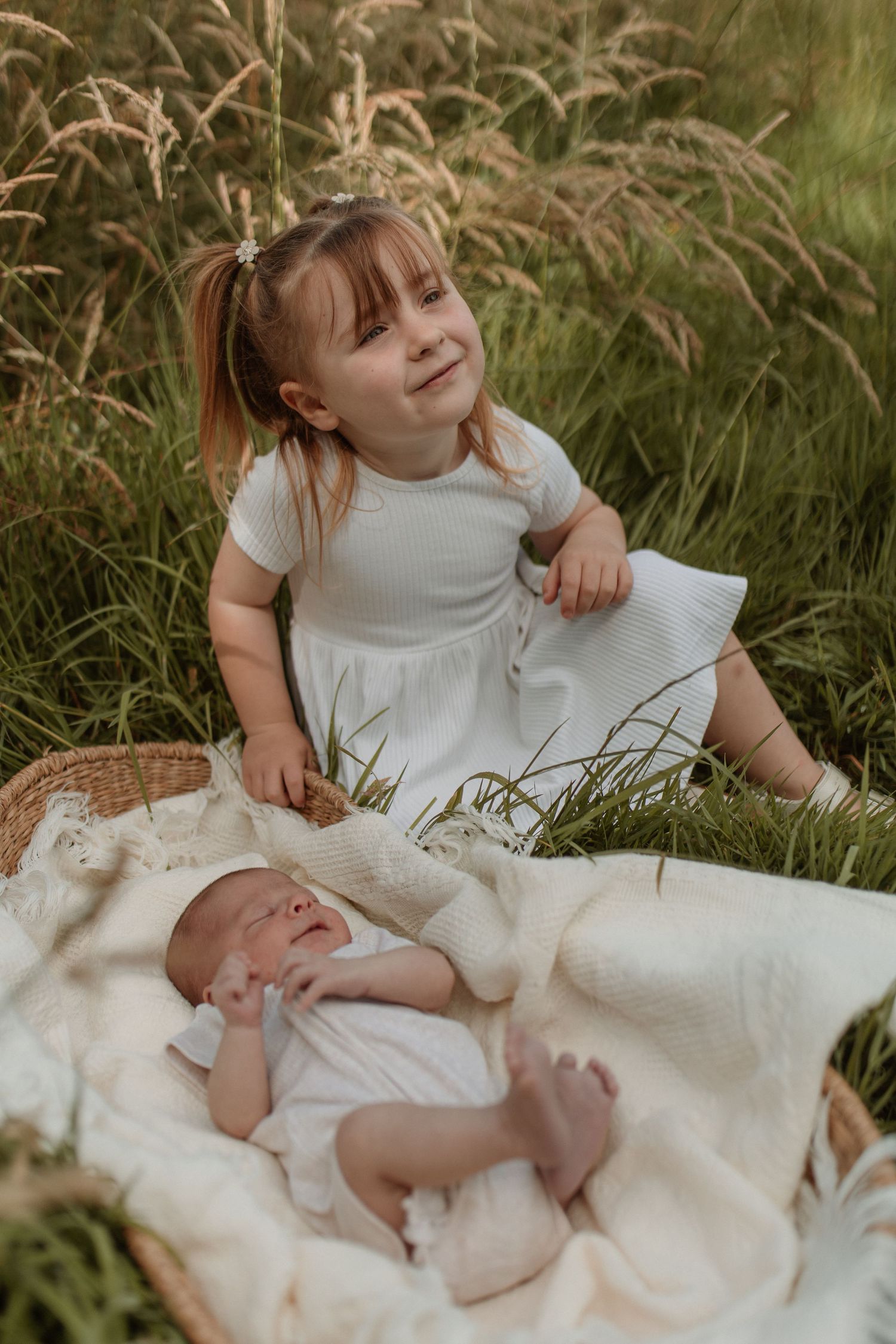 Young girl in white dress sitting in grass field beside newborn baby in basket.