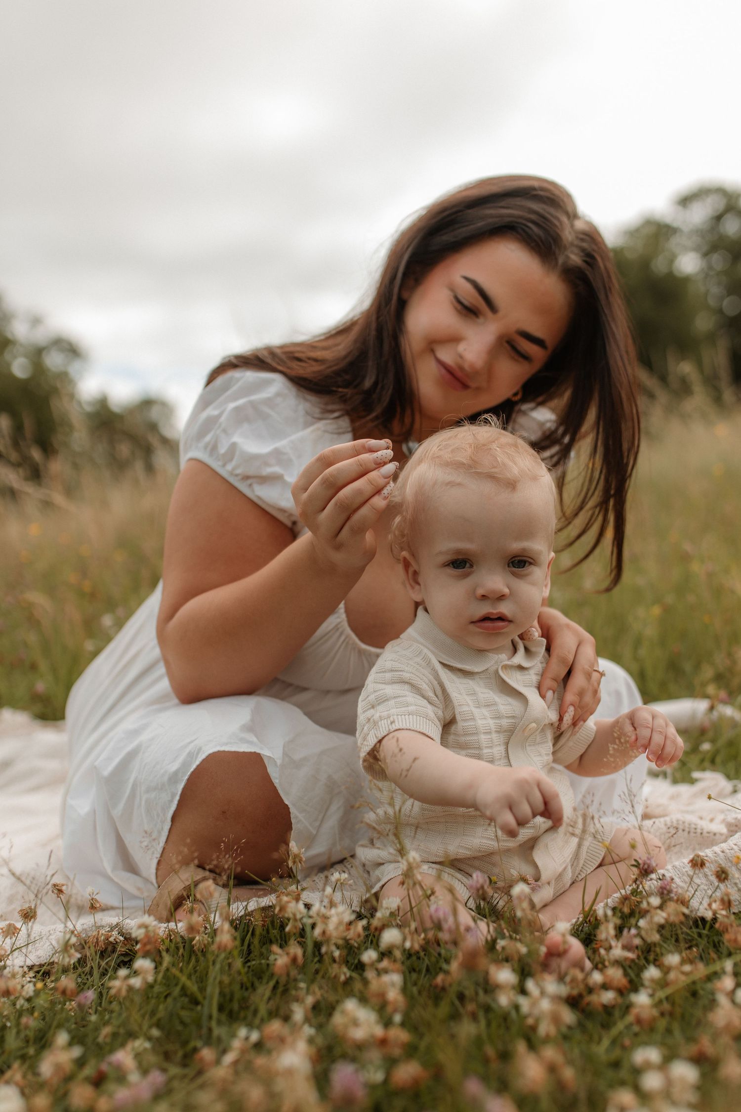 A smiling mother in a white dress sitting in a wildflower field with her baby boy on a blanket.