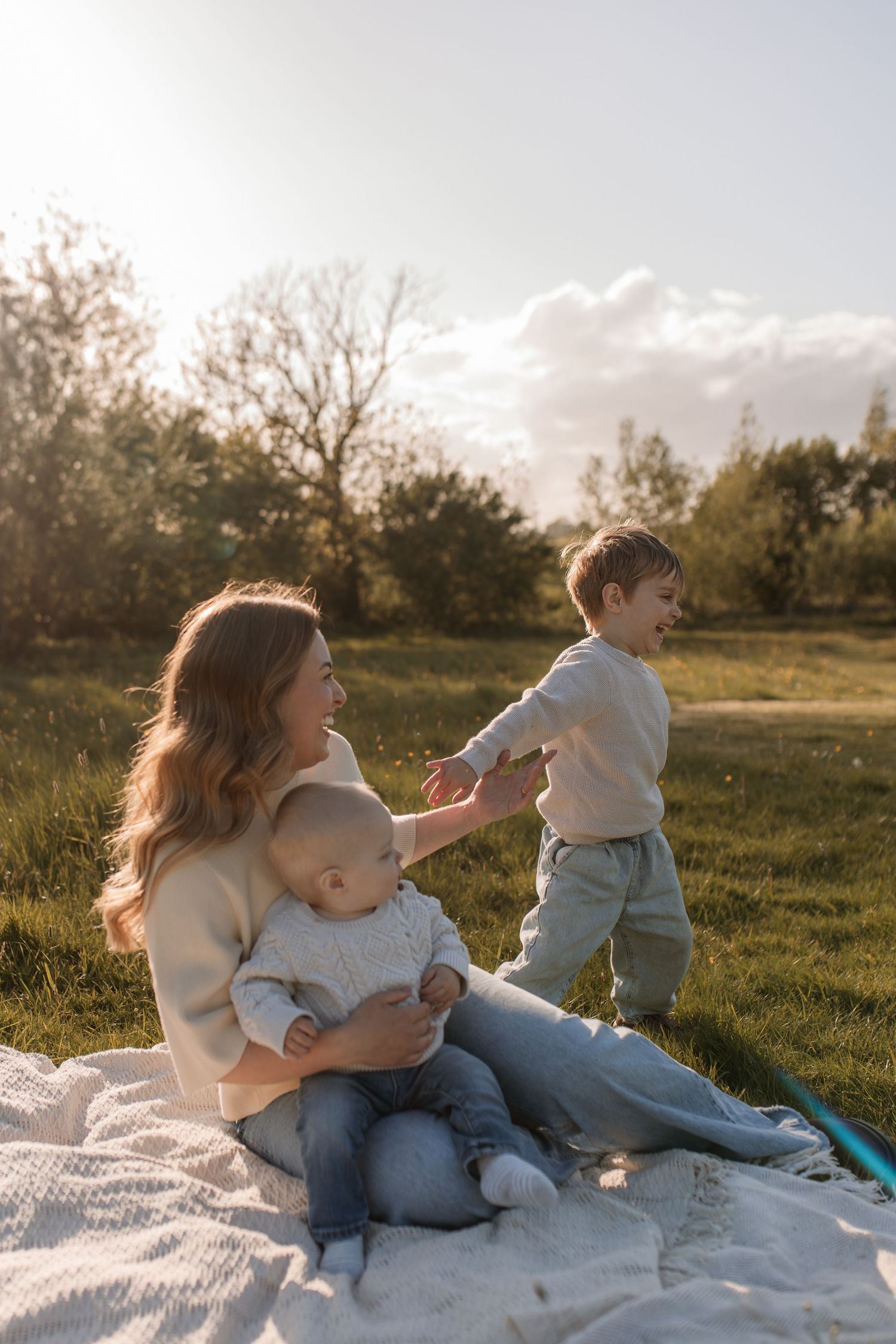 A mother sitting on a blanket in a sunny park with her baby and toddler son.