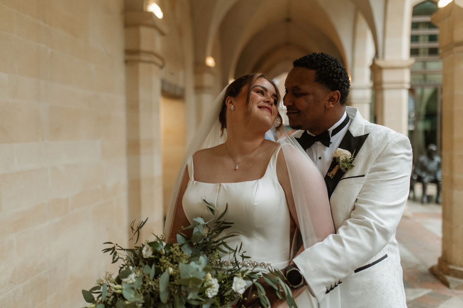 Interracial newlywed couple embracing under stone archway, bride holding lush greenery bouquet in white gown.