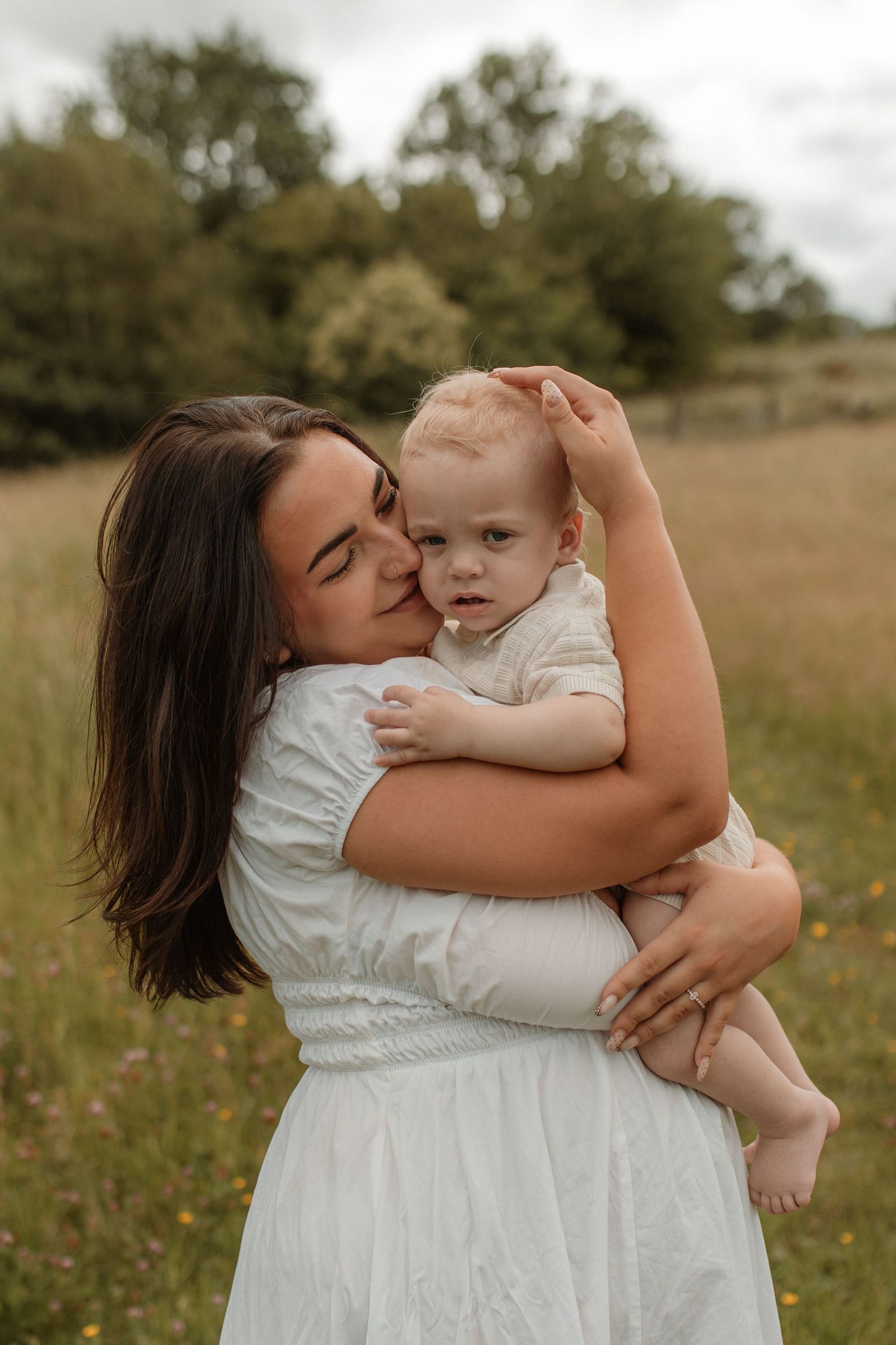 A dark-haired mother in a white dress kissing her blonde baby boy while standing in a green meadow.