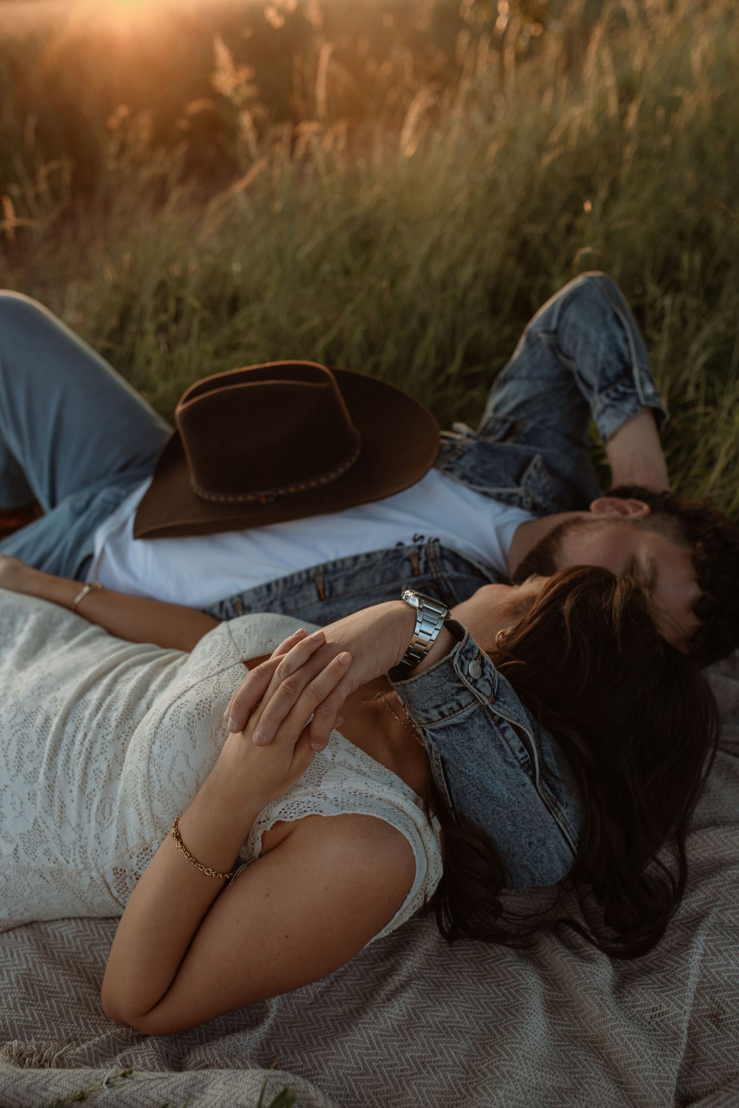 Couple lying in golden grass field at sunset, woman in white lace dress and man in denim jacket with brown hat.