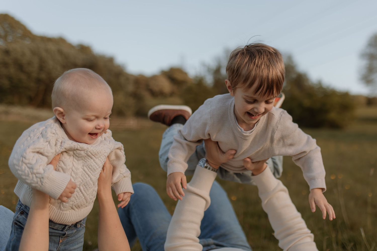 Two laughing children in cream outfits play outdoors as parents lift them up in a sunny field.