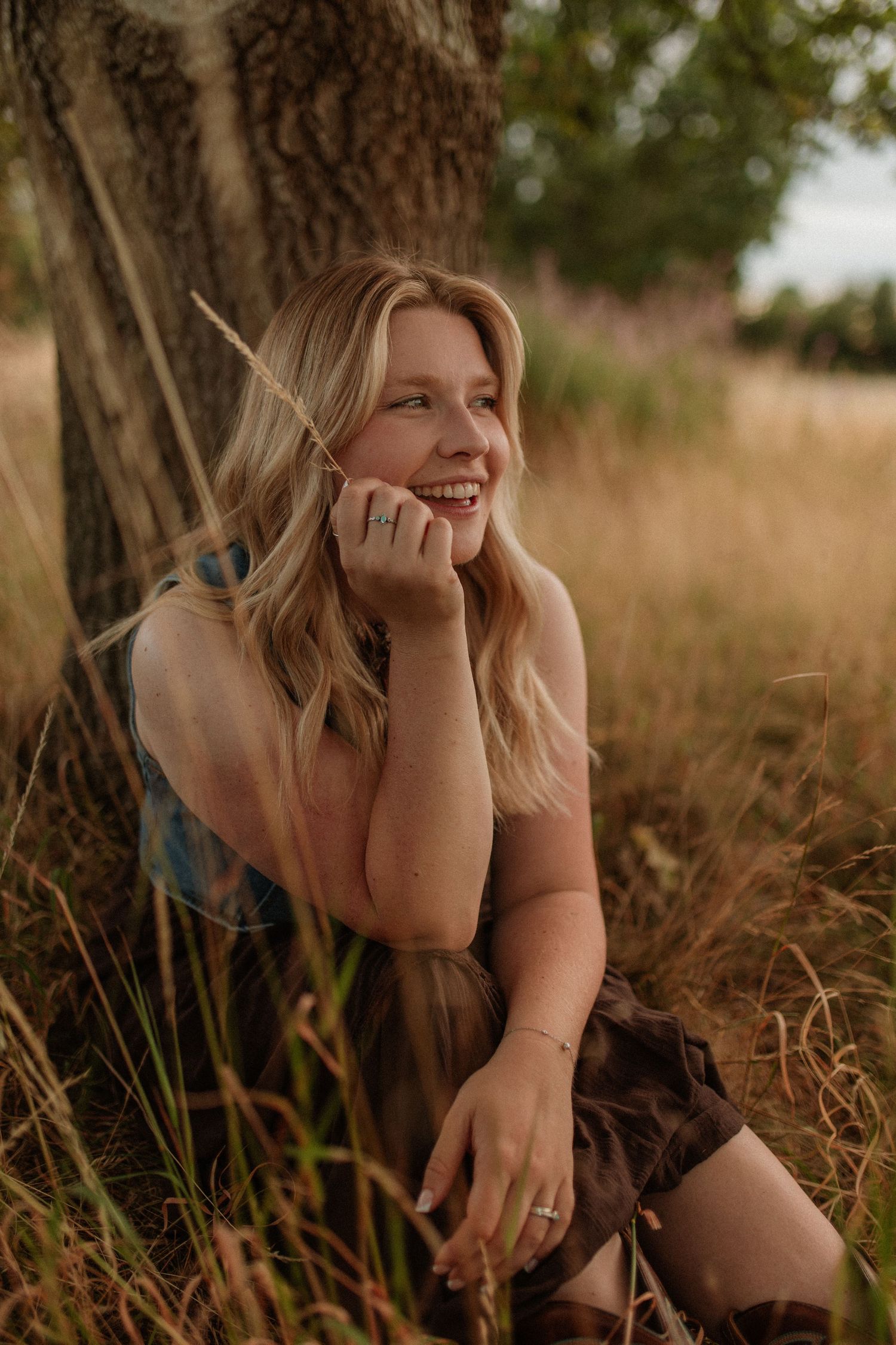 A smiling blonde woman sitting against a tree in a golden grass field outdoors.