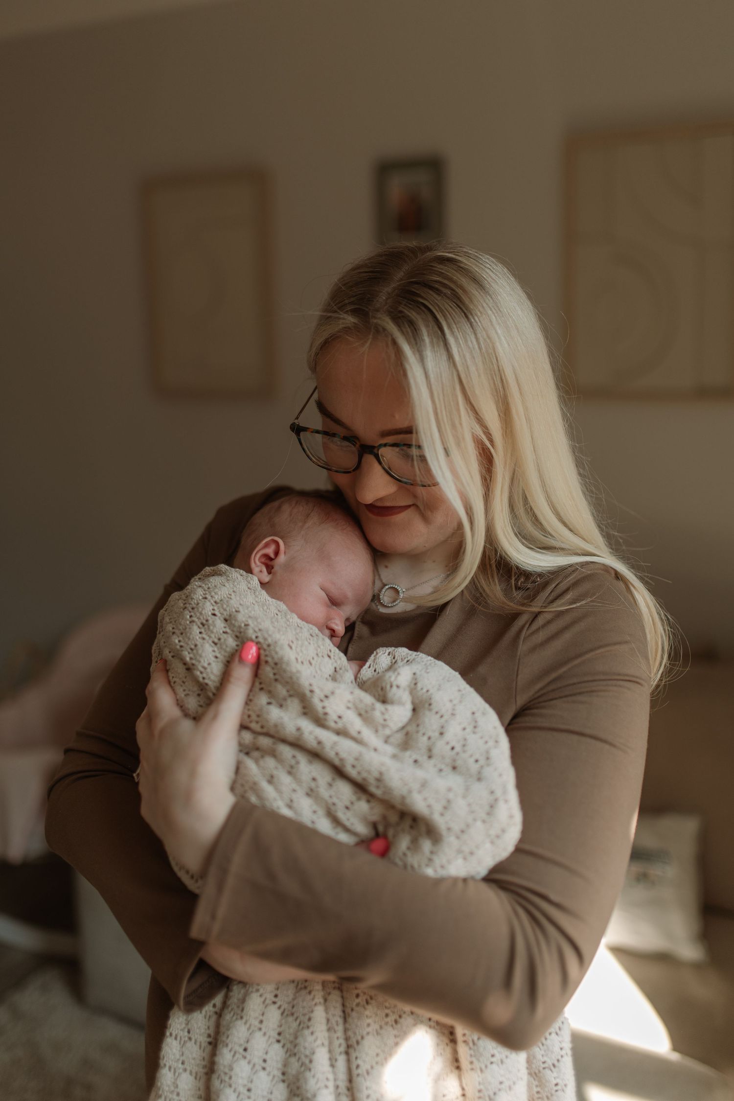 Blonde mother with glasses tenderly holding her sleeping newborn in warm indoor light.