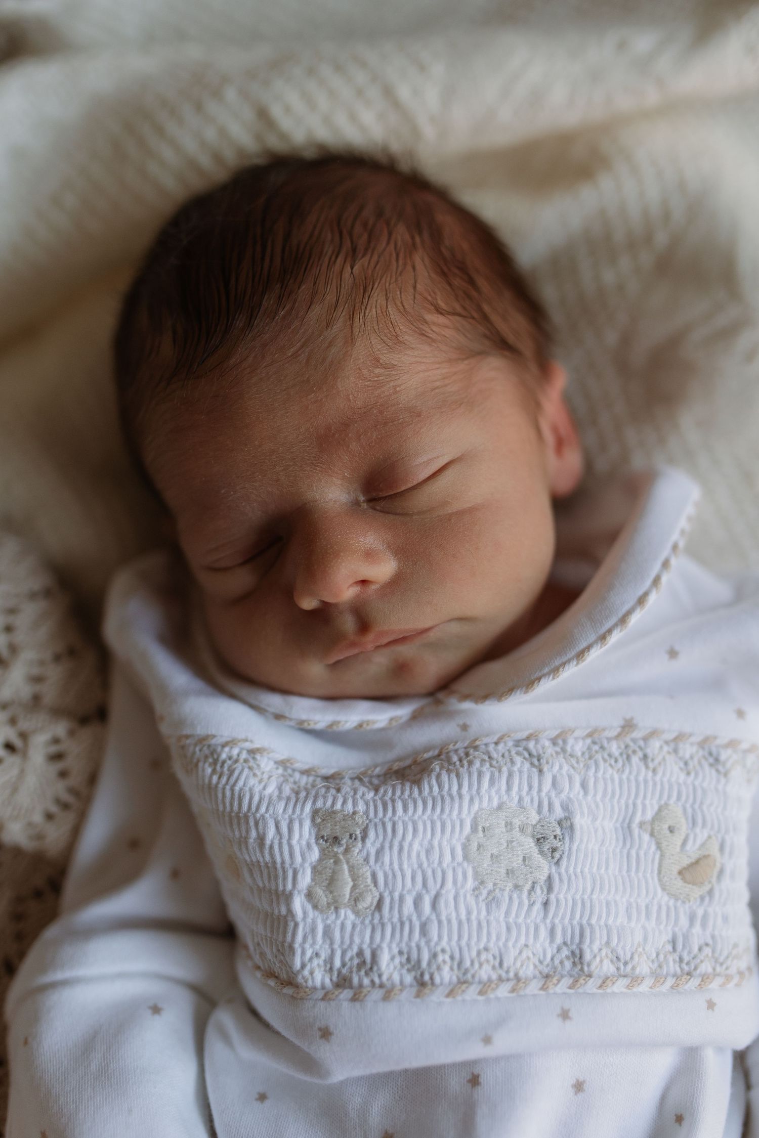 Sleeping newborn baby in white smocked outfit with teddy bear embroidery on lace blanket.