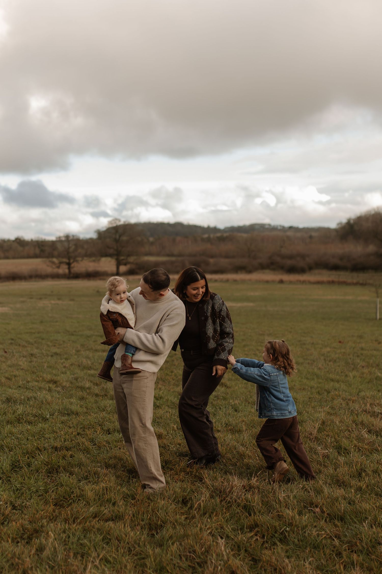 Family of four playing joyfully in an open green field on a cloudy autumn day.