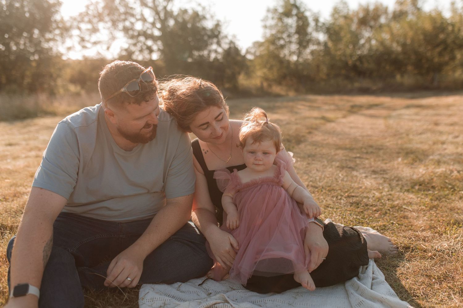 A family of three sits on a blanket in a sunny grassy field.