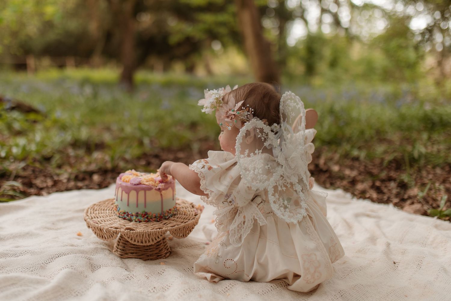 A baby girl in a white lace fairy dress reaches toward a birthday cake during an outdoor smash cake session.