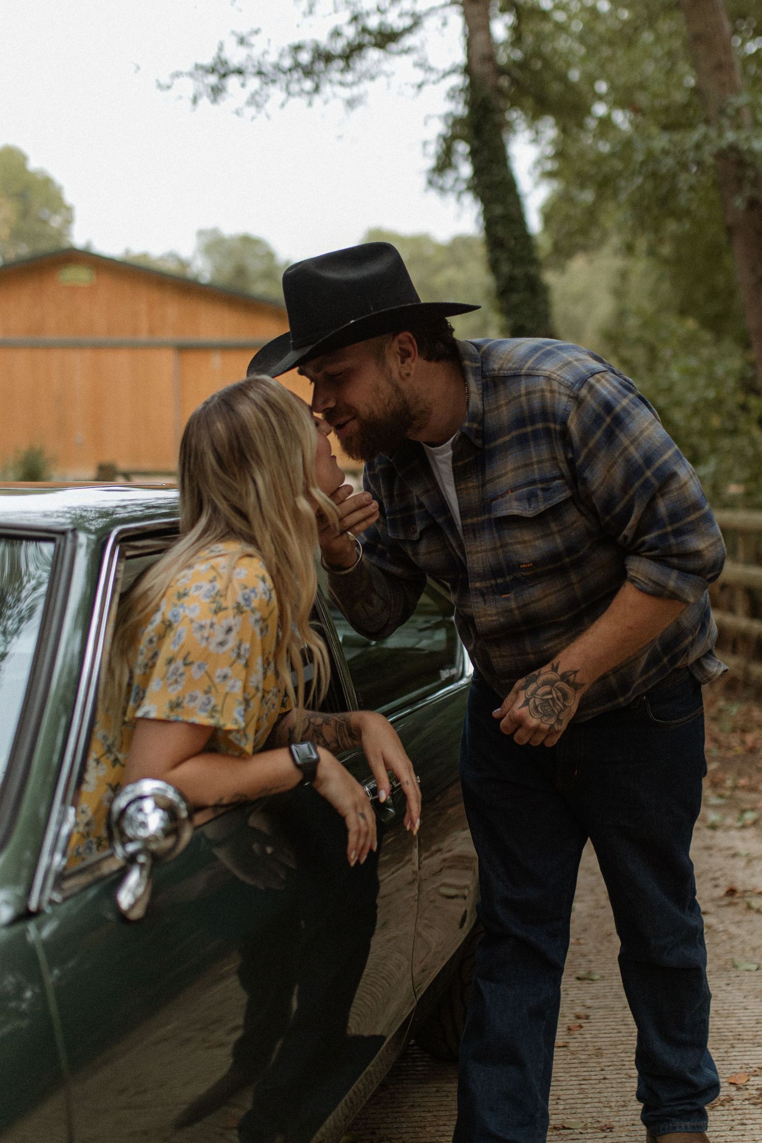 Couple shares an intimate moment leaning against a vintage green car surrounded by trees.