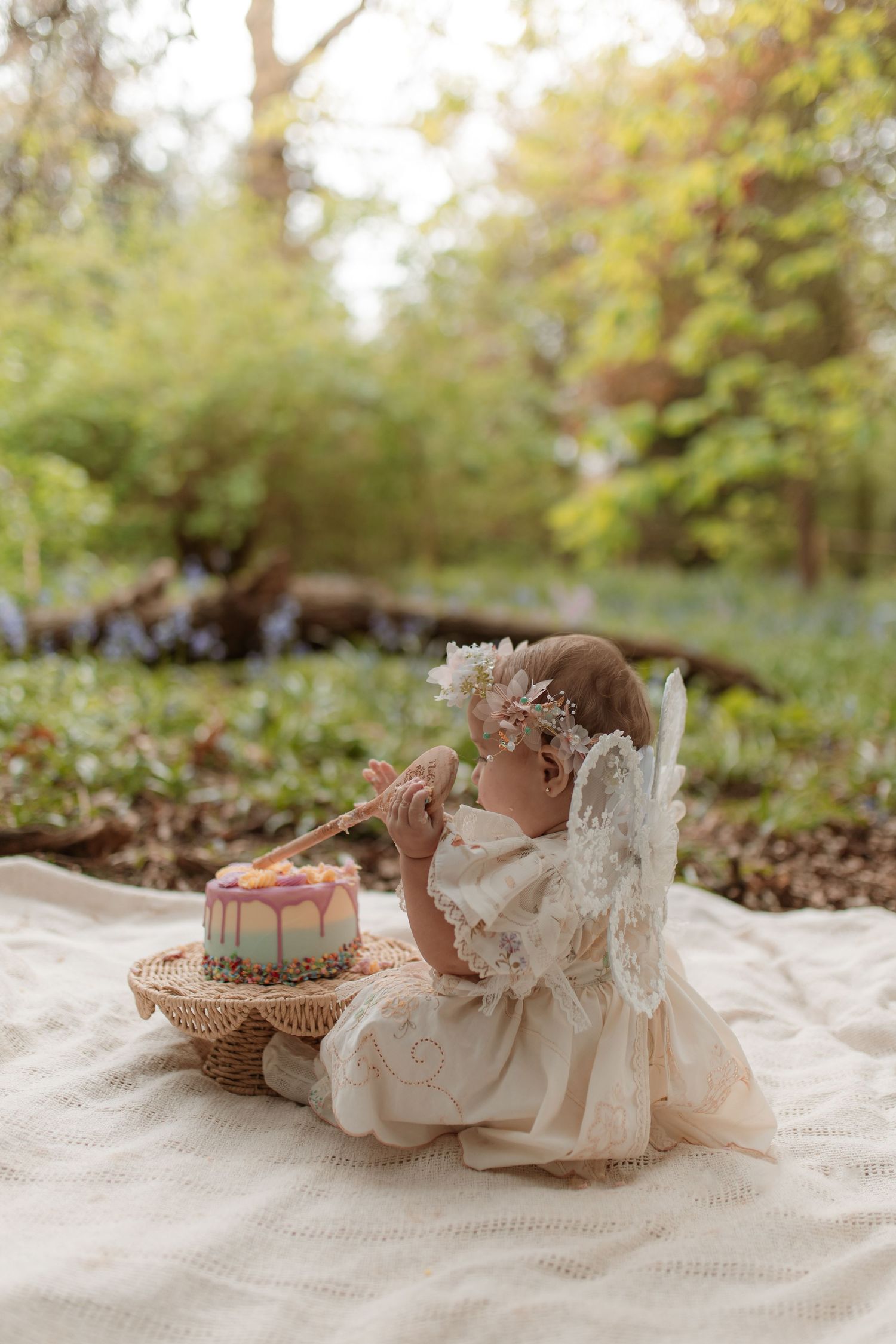 Adorable baby girl in fairy costume sitting on blanket outdoors with birthday cake in forest.